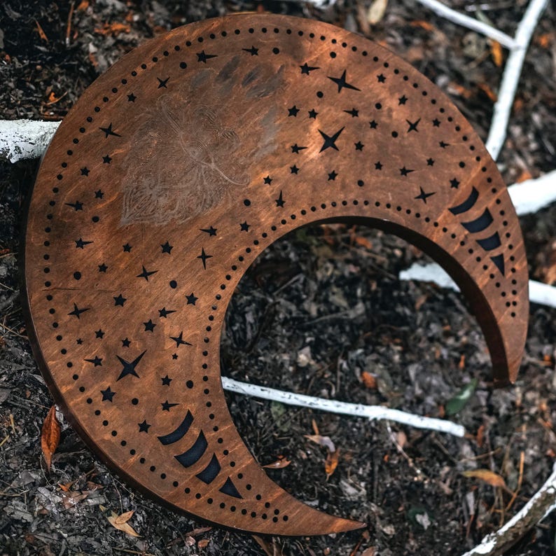 May include: A crescent moon-shaped wooden decorative piece with a dark brown finish. The moon features cut-out star and dot patterns, and a subtle etched design. The piece is resting on a bed of leaves and twigs.