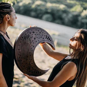 May include: A wooden crescent moon with star and crescent cutouts is held by two people. The moon is a dark brown color and has a decorative design. The people are wearing black tops.