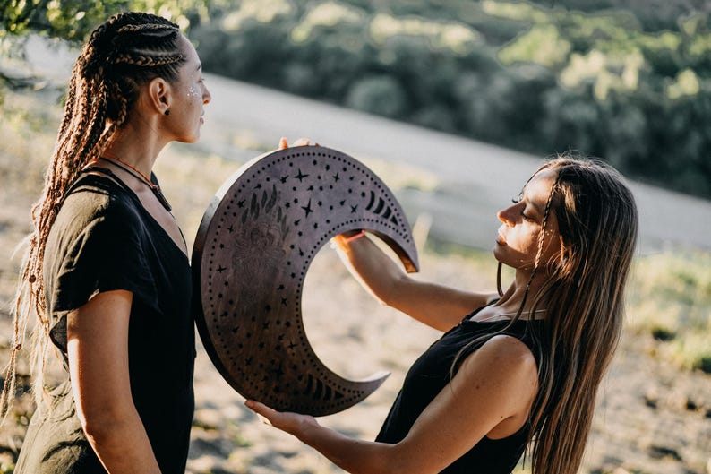May include: A wooden crescent moon with star and crescent cutouts is held by two people. The moon is a dark brown color and has a decorative design. The people are wearing black tops.