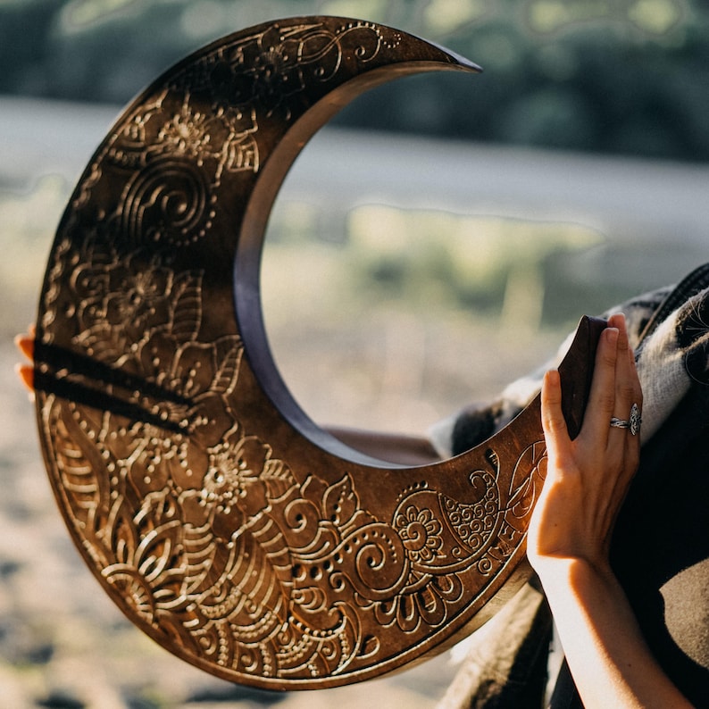 May include: A large, crescent-shaped wooden wall hanging with intricate floral and paisley carvings. The dark brown piece is held by a person, showcasing the detailed design and texture. The artwork is a decorative piece.
