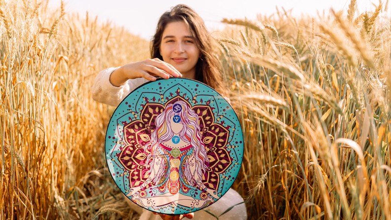 May include: A woman holds a round, turquoise-coloured mandala with a colourful illustration of a woman in a meditative pose. The mandala features a floral design with gold accents and a symbol of the third eye chakra.