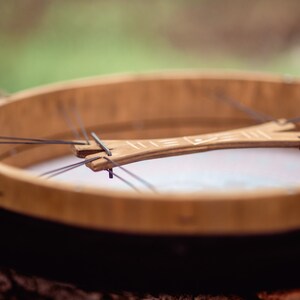 May include: A wooden frame drum with a black rim and a white leather head. The drum has a wooden crossbar with a carved design.