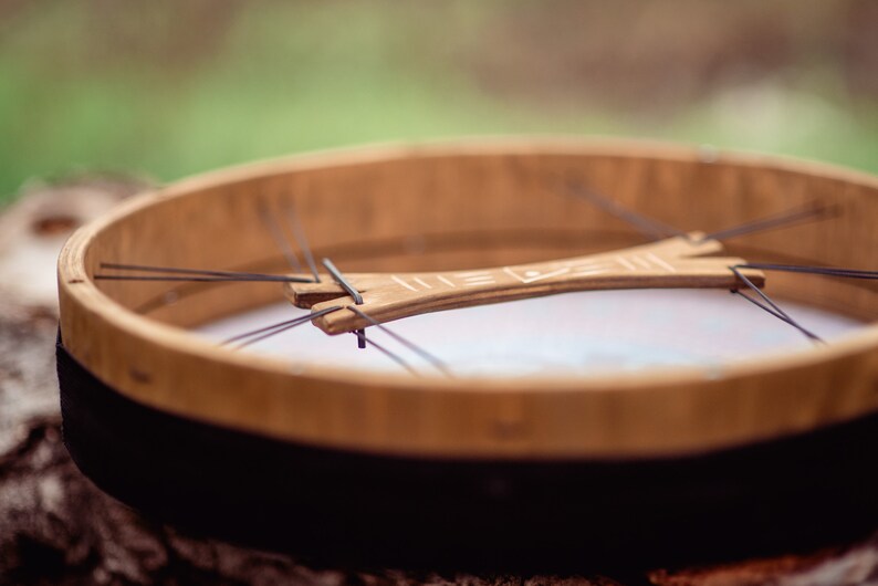 May include: A wooden frame drum with a black rim and a white leather head. The drum has a wooden crossbar with a carved design.