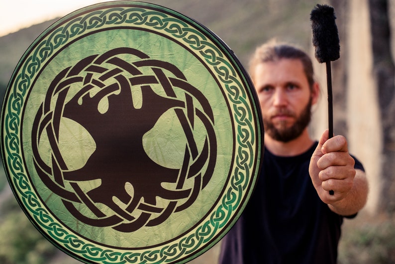 May include: A person holds a green and black circular drum with a Celtic knot design and a black stick with a fuzzy end.