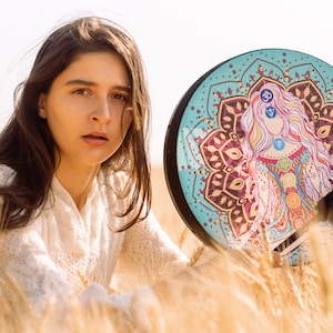 May include: A woman with long brown hair sits in a field of wheat, holding a round, turquoise-coloured drum with a mandala design featuring a woman with colourful chakras.