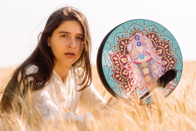 May include: A woman with long brown hair sits in a field of wheat, holding a round, turquoise-coloured drum with a mandala design featuring a woman with colourful chakras.