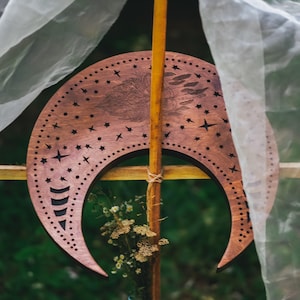 May include: A wooden crescent moon with star cutouts and a detailed engraved design. The brown moon is mounted on a wooden pole and decorated with small flowers. Sheer white fabric drapes in the background.