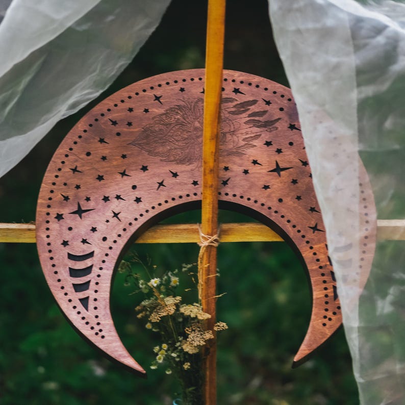 May include: A wooden crescent moon with star cutouts and a detailed engraved design. The brown moon is mounted on a wooden pole and decorated with small flowers. Sheer white fabric drapes in the background.