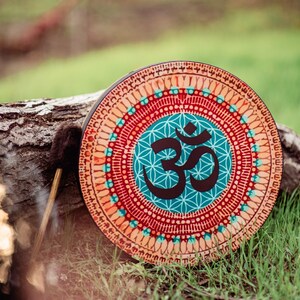 May include: A round wooden frame drum with a colourful mandala design featuring the Om symbol in the centre. The drum is resting on a log with a brown drumstick beside it.