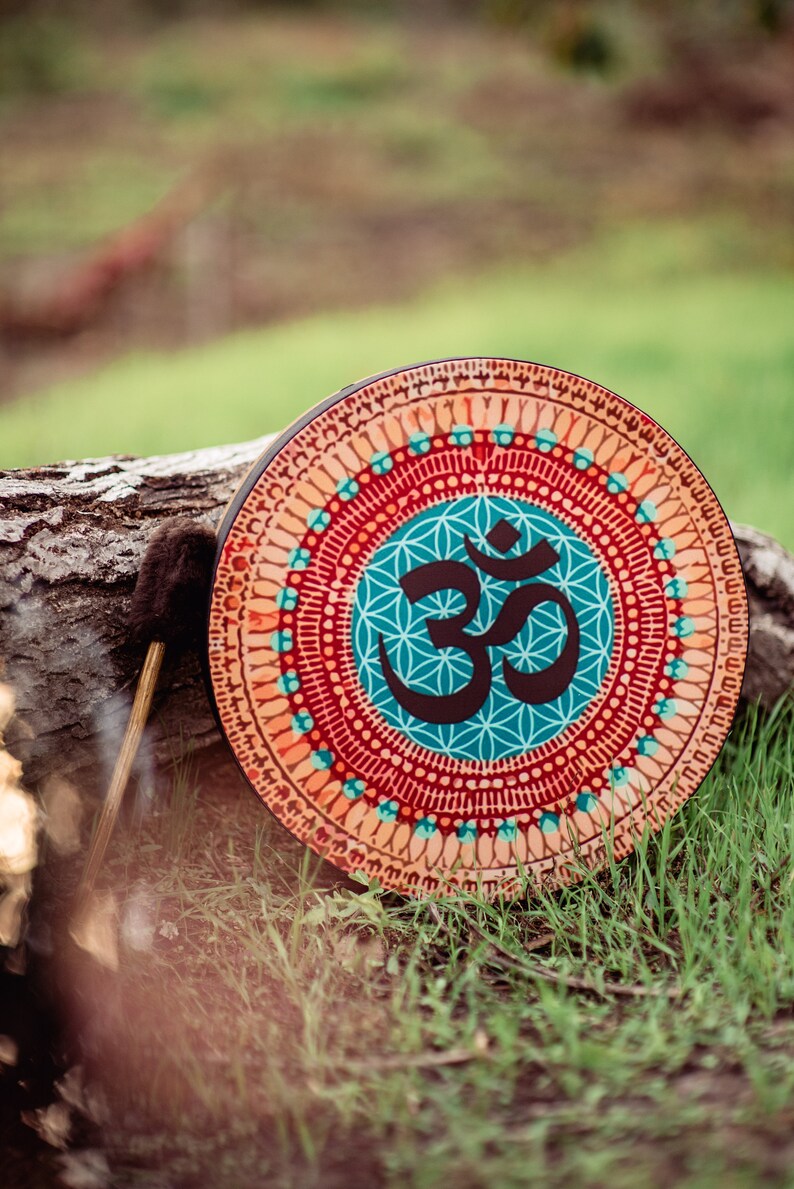 May include: A round wooden frame drum with a colourful mandala design featuring the Om symbol in the centre. The drum is resting on a log with a brown drumstick beside it.
