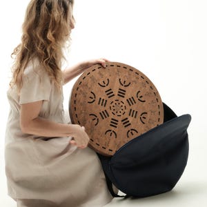 Könnte beinhalten: Ein rundes Holzobjekt mit einem geschnitzten geometrischen Design wird in eine schwarze Tragetasche gelegt. Das Holzobjekt hat eine hellbraune Farbe und eine Reihe von ausgeschnittenen Formen. Die Tragetasche ist dunkelblau.