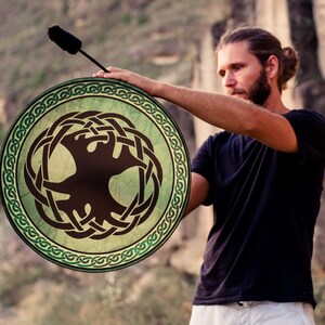 K&ouml;nnte beinhalten: Eine Person spielt eine gr&uuml;ne und braune Rahmtrommel mit einem keltischen Knotendesign und einem Baum-des-Lebens-Symbol. Die Trommel wird mit einem braunen Schl&auml;gel gespielt.