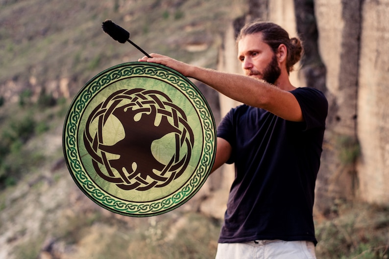 K&ouml;nnte beinhalten: Eine Person spielt eine gr&uuml;ne und braune Rahmtrommel mit einem keltischen Knotendesign und einem Baum-des-Lebens-Symbol. Die Trommel wird mit einem braunen Schl&auml;gel gespielt.