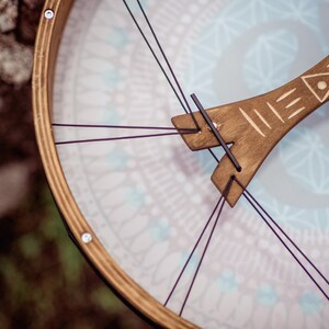 May include: A wooden frame drum with a light blue and white patterned surface. The drum has a wooden handle with a carved design and is held together with purple strings.