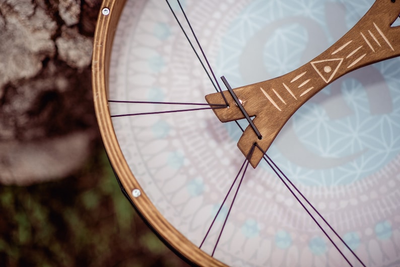 May include: A wooden frame drum with a light blue and white patterned surface. The drum has a wooden handle with a carved design and is held together with purple strings.