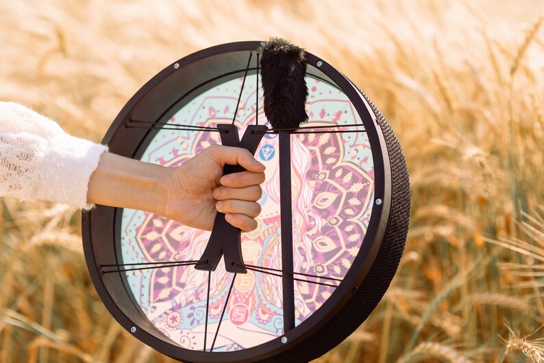 May include: A black frame drum with a colourful mandala design and a black handle. The drum is being held by a person's hand.