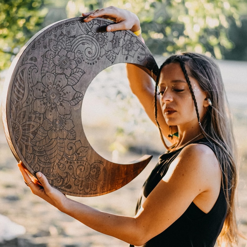 May include: A crescent moon-shaped wooden wall hanging with intricate floral and paisley carvings. The dark wood contrasts with the detailed, light-colored etched designs. The artwork is held up in front of a blurred background.