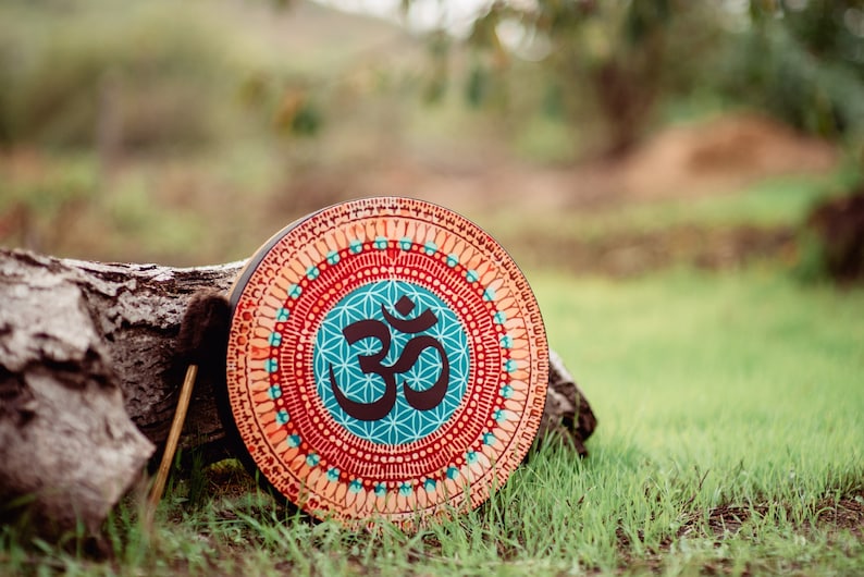 May include: A round wooden frame drum with a colourful mandala design featuring the Om symbol in the centre. The drum is resting on a tree trunk in a grassy field.