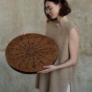 May include: A circular wooden board featuring a dark brown finish with a sunburst pattern of etched lines radiating from the center. The outer edge is decorated with a repeating geometric triangle border, and the wood grain is visible throughout.