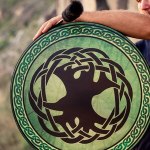 K&ouml;nnte beinhalten: Eine gr&uuml;ne und schwarze Handtrommel mit einem keltischen Knotendesign und einem Lebensbaummotiv. Die Trommel wird von einer Person gehalten.