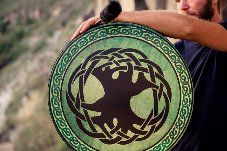 K&ouml;nnte beinhalten: Eine gr&uuml;ne und schwarze Handtrommel mit einem keltischen Knotendesign und einem Lebensbaummotiv. Die Trommel wird von einer Person gehalten.