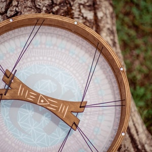 May include: A wooden frame drum with a light blue and pink patterned head. The drum is strung with black cords and has a wooden tuning key.