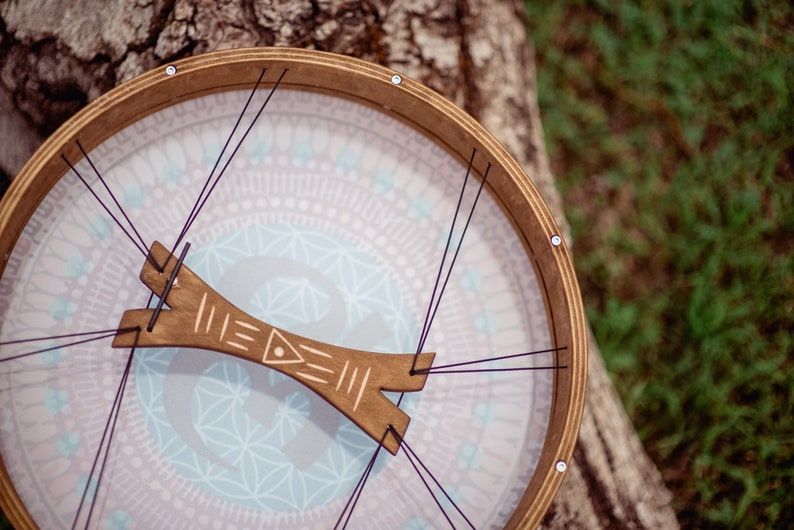 May include: A wooden frame drum with a light blue and pink patterned head. The drum is strung with black cords and has a wooden tuning key.