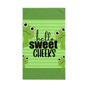 May include: A green and white striped towel featuring four cartoon frogs and the text "hello sweet CHEEKS" in black. The frogs have white eyes and are smiling. The towel has a solid green border.