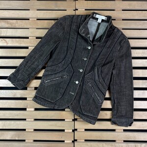 May include: A dark grey denim jacket with a stand-up collar, button closure, and multiple pockets with zips. The jacket has decorative stitching and a slim fit.