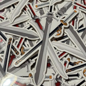 Könnte beinhalten: Eine Nahaufnahme eines Stapels von Aufklebern mit verschiedenen Schwertmustern. Die Schwerter sind silber und schwarz mit roten Akzenten. Einige der Schwerter haben Schädelmuster auf den Klingen.