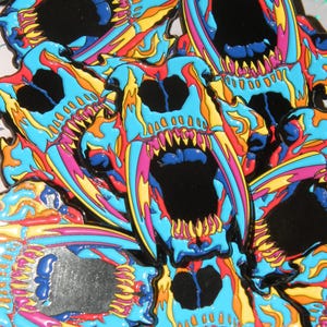 Puede incluir: Primer plano de varios pines de esmalte que presentan una calavera estilizada de colores con dientes afilados y un esquema de colores azul brillante, rosa, amarillo y naranja.
