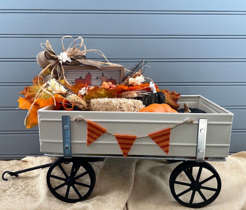 May include: A white metal wagon filled with fall decor, including pumpkins, gourds, and fall leaves. The wagon has black wheels and a wooden sign with the words "Fall" on it.