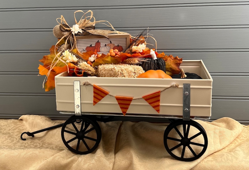 May include: A white metal wagon filled with fall decor, including faux pumpkins, hay bales, and fall leaves. The wagon has black wheels and a banner with orange and white stripes.
