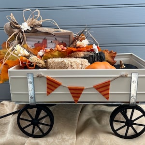 May include: A white metal wagon filled with fall decor, including pumpkins, gourds, hay bales, and fall leaves. A small wooden sign with the text "Pumpkin Patch" is visible in the wagon. The wagon has black wheels and a metal handle.