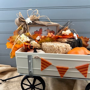 May include: A white metal wagon filled with fall decor, including faux pumpkins, hay bales, and fall leaves. A banner with orange and red stripes hangs from the wagon.