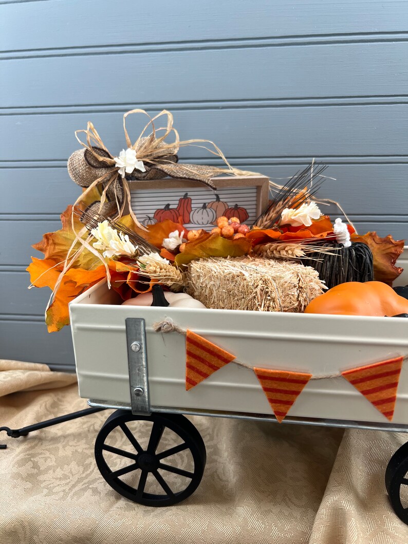 May include: A white metal wagon filled with fall decor, including faux pumpkins, hay bales, and fall leaves. A banner with orange and red stripes hangs from the wagon.