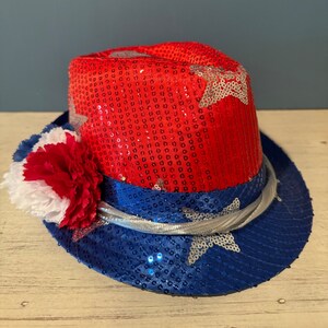 May include: A red and blue sequined fedora hat with a white star design. The hat has a white and red carnation flower on the side.