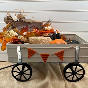 May include: A white metal wagon filled with fall decor, including pumpkins, hay bales, and fall leaves. A banner with orange and white stripes hangs across the front of the wagon.