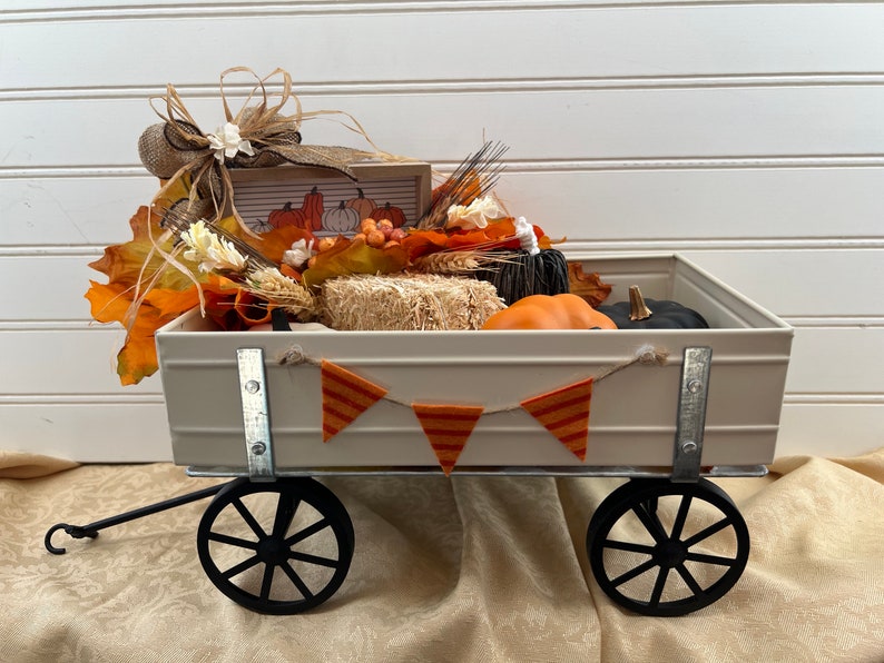 May include: A white metal wagon filled with fall decor, including pumpkins, hay bales, and fall leaves. A banner with orange and white stripes hangs across the front of the wagon.