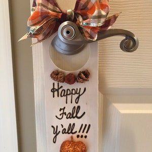 May include: A white wooden door hanger with the text "Happy Fall y'all!!!" written in black marker. The hanger is decorated with a brown and orange plaid bow, pine cones, and a glittery orange pumpkin.