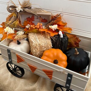May include: A white metal wagon filled with fall decor, including pumpkins, hay bales, and fall leaves. The wagon has black wheels and a banner with orange and white triangles.