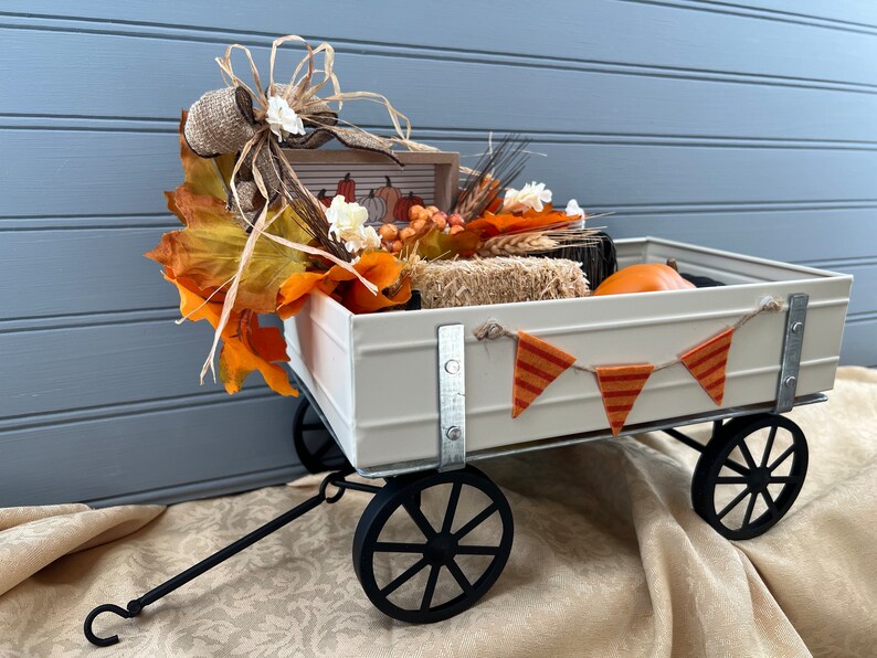 May include: A white metal wagon with black wheels and a black handle. The wagon is filled with fall decorations, including a burlap bow, orange and yellow fall leaves, a small sign with pumpkins, and a small orange pumpkin.