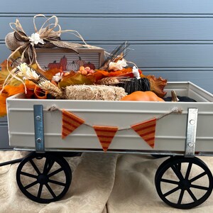 May include: A white metal wagon filled with fall decor, including pumpkins, gourds, hay bales, and fall leaves. A burlap bow with a white flower is on top of the wagon. A small sign with the text "Fall" is in the wagon.