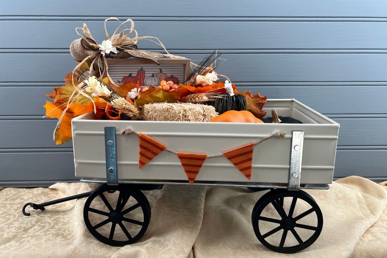May include: A white metal wagon filled with fall decor, including pumpkins, gourds, hay bales, and fall leaves. A burlap bow with a white flower is on top of the wagon. A small sign with the text "Fall" is in the wagon.