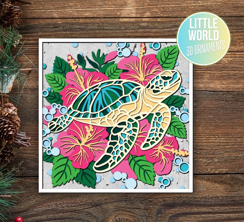 Op de afbeelding: Een 3D-papierknutsel ornament van een zeeschildpad omringd door roze hibiscus bloemen en groene bladeren. De schildpad is geel met een blauwe en groene schild. Het ornament is op een witte achtergrond.