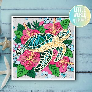Op de afbeelding: Een 3D-papierkunsthanger met een zeeschildpad omringd door roze hibiscusbloemen. De schildpad is gemaakt van gelaagd papier en heeft een lichtblauwe en groene schild. De bloemen zijn ook gemaakt van gelaagd papier en hebben een roze en witte kleurstelling. De hanger maakt deel uit van de "Little World 3D Ornaments" collectie.