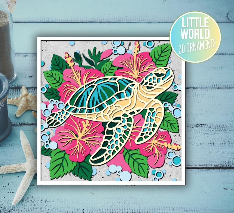 Op de afbeelding: Een 3D-papierkunsthanger met een zeeschildpad omringd door roze hibiscusbloemen. De schildpad is gemaakt van gelaagd papier en heeft een lichtblauwe en groene schild. De bloemen zijn ook gemaakt van gelaagd papier en hebben een roze en witte kleurstelling. De hanger maakt deel uit van de "Little World 3D Ornaments" collectie.