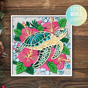 Op de afbeelding: Een 3D-papierknutsel ornament van een zeeschildpad omringd door roze hibiscus bloemen en groene bladeren. Het ornament is ontworpen om te worden geassembleerd en versierd.