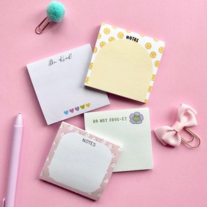 Op de afbeelding: Vier verschillende plakbriefjes in pastelkleuren met verschillende ontwerpen. De ontwerpen omvatten een gele bloc met smileygezichten, een roze bloc met witte madeliefjes, een witte bloc met hartjes en een groene bloc met een kikker en een bloem. De tekst "NOTES" staat op drie van de blokken. De tekst "DO NOT FROG-ET" staat op het groene bloc. De tekst "Be Kind" staat op het witte bloc met hartjes.