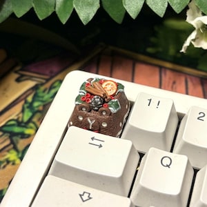 May include: A white mechanical keyboard with a custom keycap featuring a miniature cake design with berries and orange slices. The keyboard has standard letter and number keys.